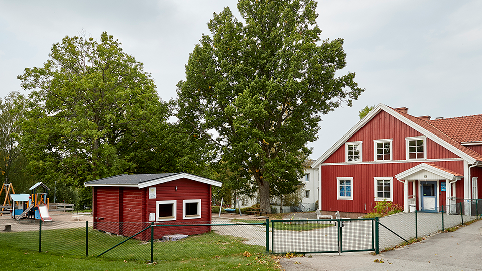 Ett rött flervåningshus i trä med vita husknutar. Framför huset en skolgård med klätterställning. 